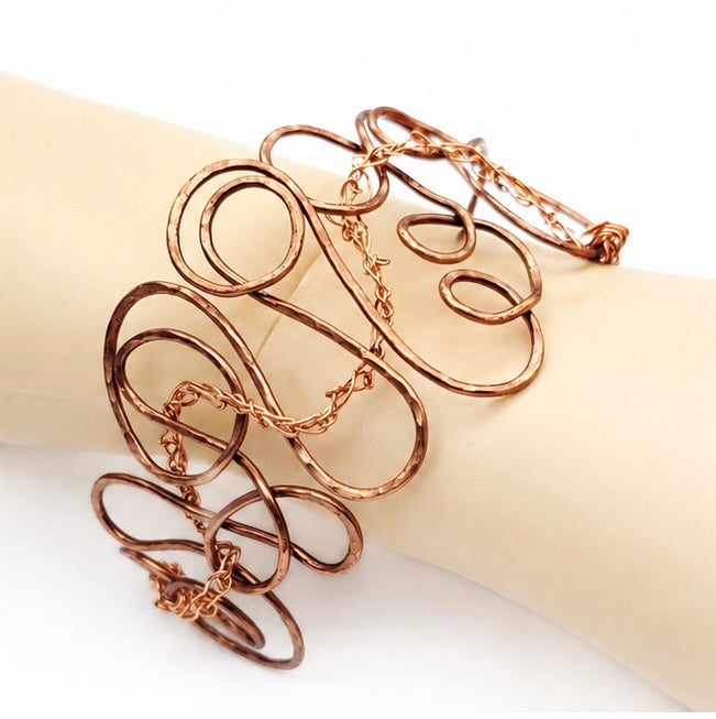 Bold antiqued copper wire bangle shown worn on a wrist, featuring open sculptural loops and woven copper details